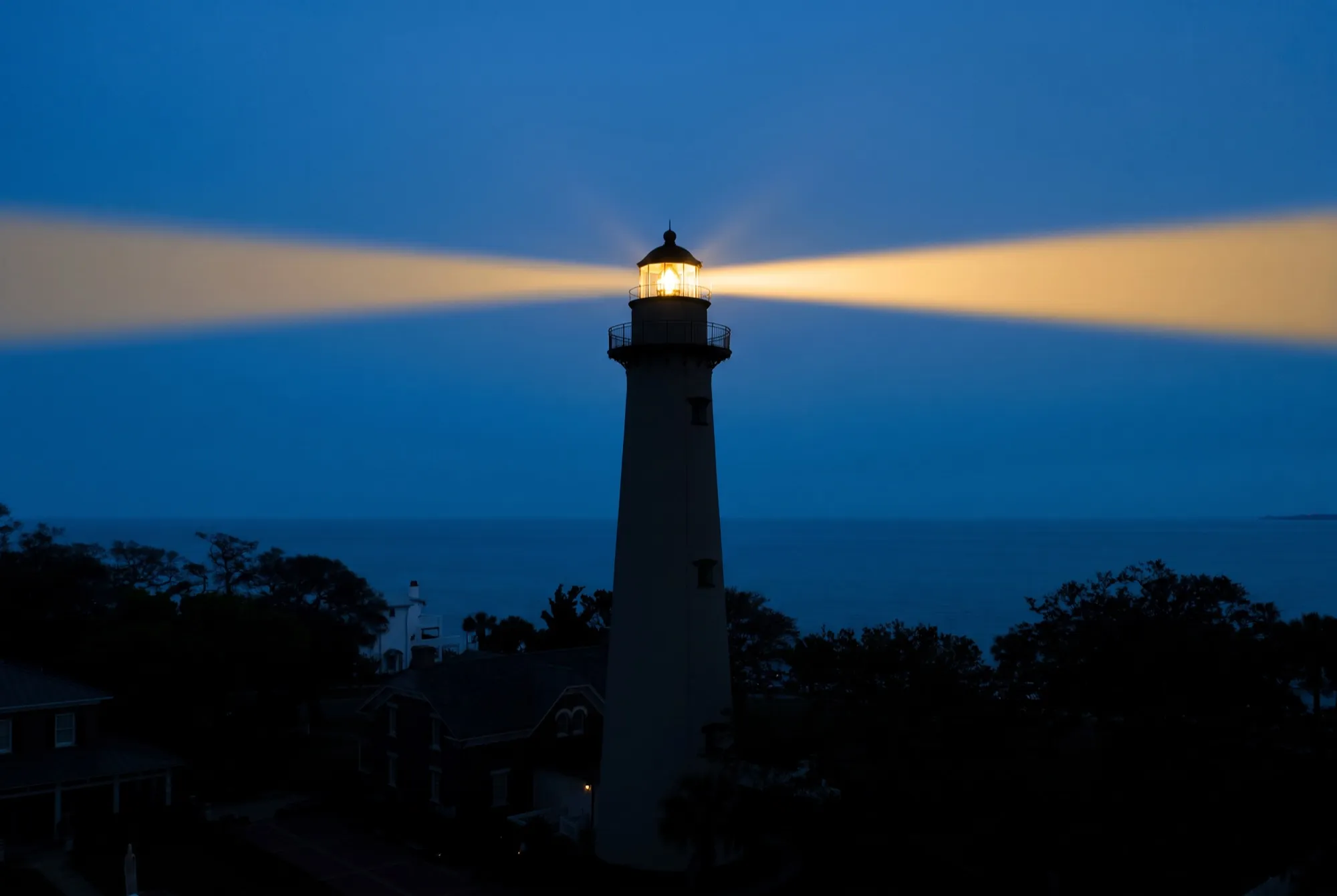The St. Simons lighthouse at dusk, its warm gold beam cutting across a deep blue sky