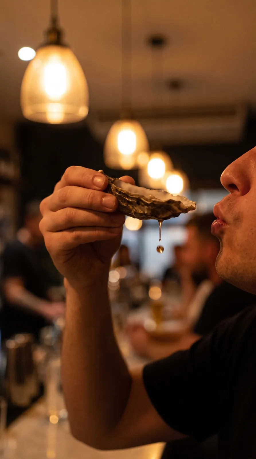Hand tipping an oyster shell toward the lips, bar bokeh behind